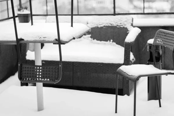 Outdoor furniture covered with snow, black and white. Snow covered chairs and sofa on balcony. Winter in the city. Town covered with fresh snow. Frozen town. Outdoor cafe in snowy winter. Snowy weather.