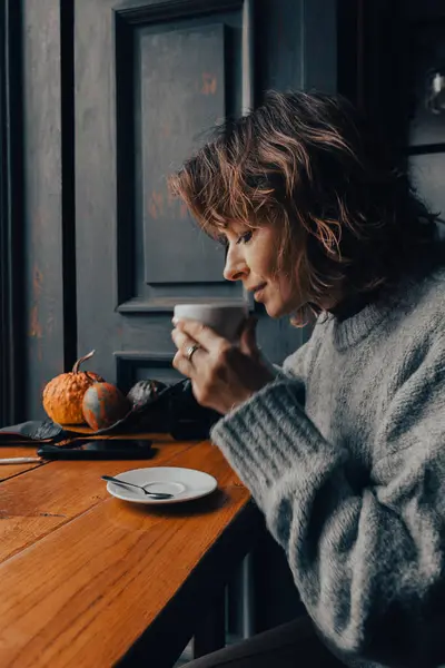 Woman in cafe with coffee. Autumn cozy cafe. Girl in knitted sweater with coffee cup and pumpkins. Beautiful girl in elegant cafe interior. Morning coffee time on cloudy autumnal day. Cozy lifestyle.