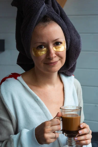 Young woman with eye patches and towel drinking morning coffee. Girl after bath with skin mask. Skin care at home. Healthy lifestyle. Pleasure procedure after taking bath. Home cosmetology.