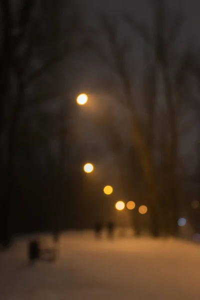 Winter park with street lamps at night, blurred. Blizzard in the city, defocused. Winter night landscape. Frozen weather background. February weather. Snow and freeze.