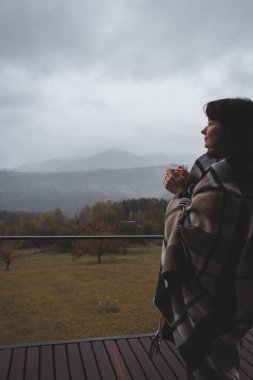 Sisli dağ manzarasında kareli ekoseli çay içen bir kız. Gezgin sonbahar dağlarında dinleniyor. Battaniyeli bir kadın kırsalda rüya görüyor. Karpatya dağlarında seyahat et. Huzur ve rahatlık. Sonbahar havası. 