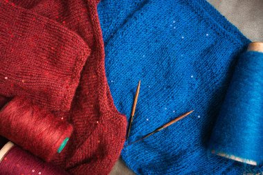 Pullu ve örgü iğneli kırmızı ve mavi süveter. Ahşap iğnelerin üzerine dizilmiş örgü giysiler. İğne işlerinde sequin deseni. Örgü hobisi. Örgü işi. Dokuları örgü kumaşla kaplıyor. Parlak örgü moda..