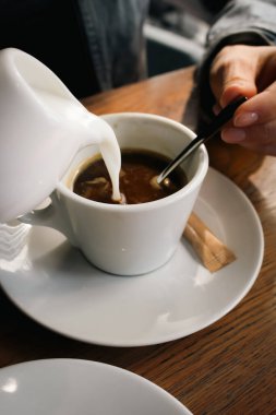 Kahve fincanına süt koyan kız. Elleri kaşıkla sütlü kahveyi karıştırıyor. Kafede Latte. Sütlü espresso. Sıcak içecek konsepti. Kahveyle rahatla. Kahvaltı konsepti. Kafeteryadaki kadın..