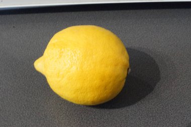 Parlak sarı bir limon zarif bir şekilde parlak bir yüzeye oturur, ışığı yakalar ve hafif desenli cildini ön plana çıkarır. Altın parıltısı tazeliği ve canlılığı çağrıştırırken, keskin aroması görüntüde neredeyse elle tutulur gibi görünüyor..