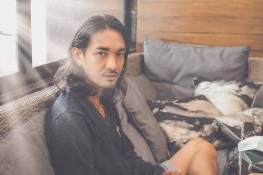 Portrait of a handsome Asian man with long hair sitting on a sofa looking at the camera with a warm glow from behind.