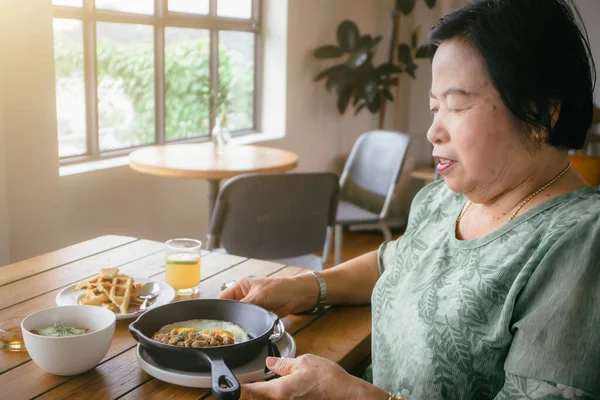 stock image An elderly Asian woman in a traditional green shirt sitting on a chair smiling happily with breakfast set on the table in a restaurant.