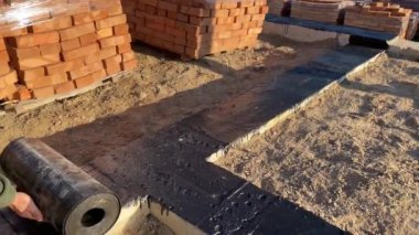 Gaz bloğundan ya da tuğladan duvar örmeye başlamadan önce binanın temelini su geçirmez hale getirin. Ağı güçlendiriyorum. Bir adam binaların tepesinde su geçirmez bir rulo yuvarlıyor.