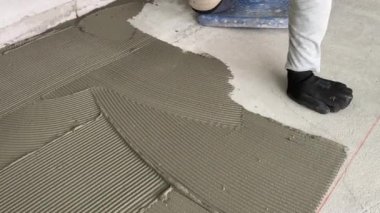 A builder applies tile adhesive to the floor. 4k video footage of construction work on laying tiles. A man using a notched trowel applies a thin layer of adhesive to the subfloor