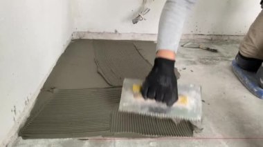A builder applies tile adhesive to the floor. 4k video footage of construction work on laying tiles. A man using a notched trowel applies a thin layer of adhesive to the subfloor