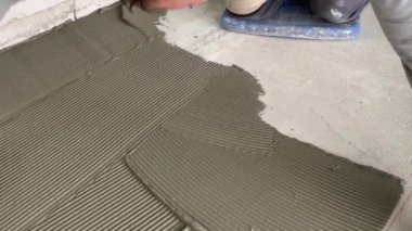 A builder applies tile adhesive to the floor. 4k video footage of construction work on laying tiles. A man using a notched trowel applies a thin layer of adhesive to the subfloor