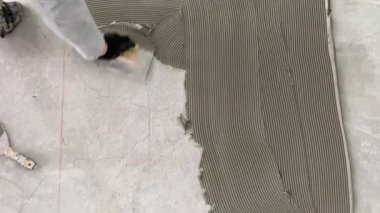 A builder applies tile adhesive to the floor. 4k video footage of construction work on laying tiles. A man using a notched trowel applies a thin layer of adhesive to the subfloor