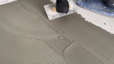 A builder applies tile adhesive to the floor. 4k video footage of construction work on laying tiles. A man using a notched trowel applies a thin layer of adhesive to the subfloor