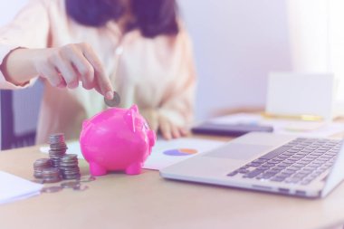 Bir kadının eli, dizüstü bilgisayarı ve mali tabloları olan pembe bir kumbaraya bozuk para atıyor..