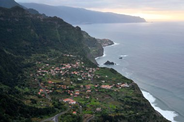 Houses of small town located on slope of mountain near waving ocean at evening time in Madeira, Portugal