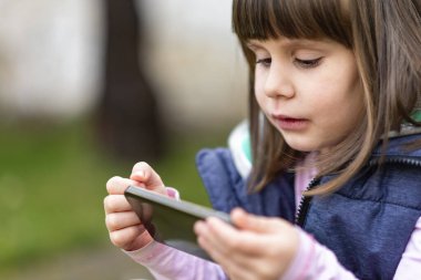 Bir elinde akıllı telefon tutan, dışarıda video izleyen, bağımlılık konsepti olan tatlı küçük bir kız.