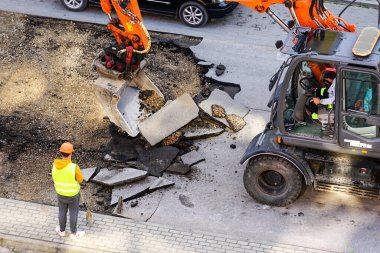 Bir kazıcı, eski asfalt tabakasını şehirdeki tamirat sırasında bir kovayla kırıyor. Kürekle çalışan bir işçi.