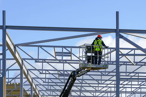 Worker in self propelled articulating boom lift basket in uniform and safety protective equipment on a building steel framework background