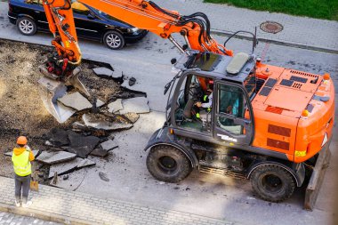 An excavator breaks up the old asphalt layer with a bucket during street repairs in city, worker with a shovel clipart