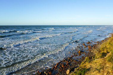 Kayalık Baltık Denizi kıyılarının güzel manzarası, kum tepesi ve beyaz köpüklü mavi dalgalar günbatımı ışığında, ufukta mavi gökyüzü