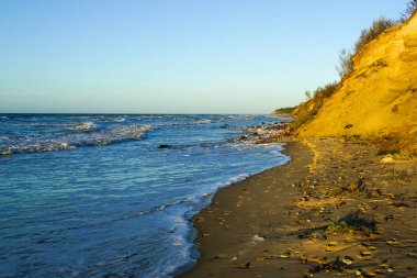 Beautiful view of rocky Baltic Sea coast, high sand dune and blue waves with white foam tops in sunset light, blue sky over horizon