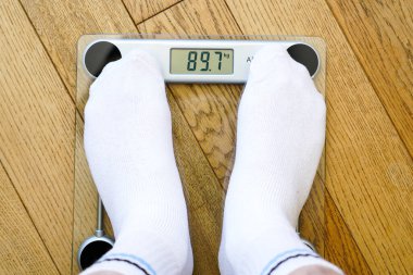 A man in white socks weighing himself on a transparent electronic floor scale, the weight reading is 89 kg, top view