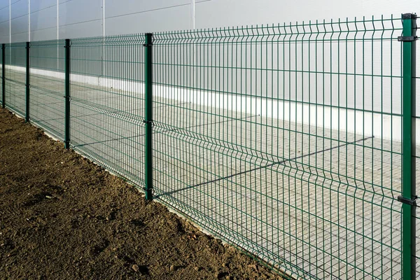 stock image A new green metal grid fence around a new industrial site, grass soil on one side and a paved lane on the other