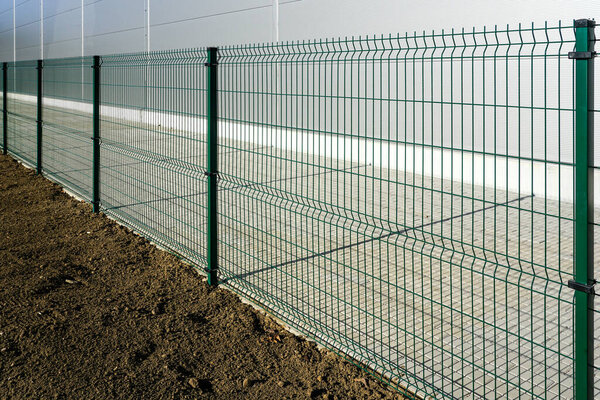 A new green metal grid fence around a new industrial site, grass soil on one side and a paved lane on the other