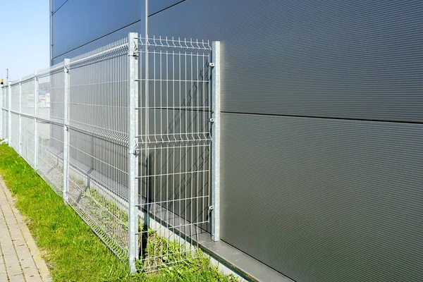 A new galvanized metal wire fence around a new gray industrial facility, green grass and a paved lane