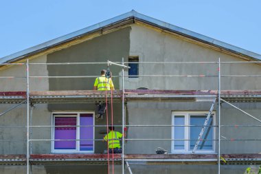 Yeşil güvenlik yelekli iki boyacı bir binanın ön cephesini iskele ve boya silindiri kullanarak boyarlar, binanın ön cephesini yeniler.