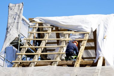 Eski ahşap bir evin hasarlı çatı yapısını, beyaz güvenlik ağını, mavi gökyüzü arka planını değiştiren bir işçi.