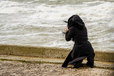 Siyah ceketli bir kadın dizlerinin üzerinde fırtınalı bir denizi telefonuyla, güçlü rüzgarla, dikiz aynasıyla fotoğraflıyor.