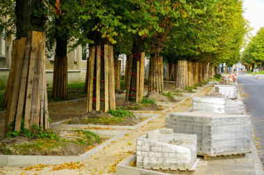 Sokak ve kaldırımlar yeniden inşa ediliyor, ağaç gövdeleri korunuyor tahta örtülü, perspektif manzaralı, paletler kaldırım taşlarıyla kaplı.