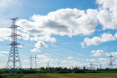 Birkaç yüksek voltajlı elektrik hattı iletim direklerine, beyaz bulutlu mavi gökyüzüne sahip endüstriyel bir manzara.
