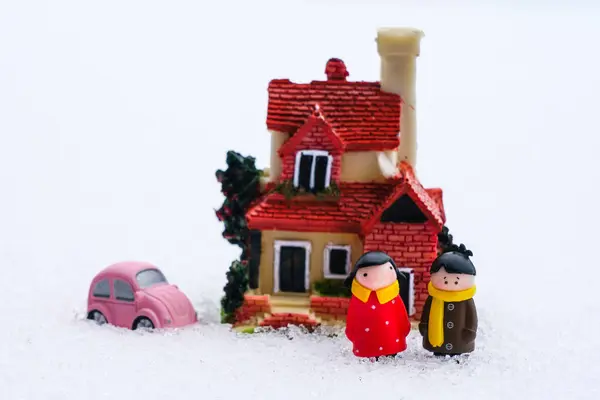 Two miniature figures of a man and a woman in the snow near a miniature residential house and little pink car, new house cost concept