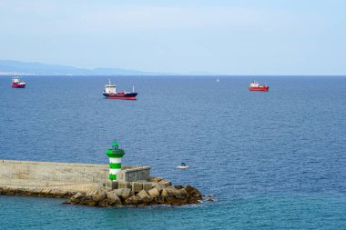 Limana yakın kargo gemisi trafiği, limanın beton iskelesi ön planda küçük yeşil bir deniz feneri, denizde nakliye.