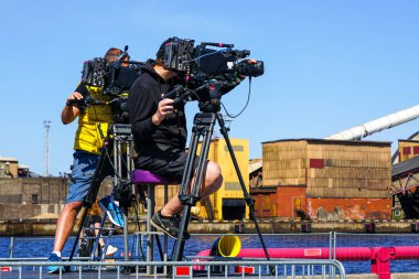 Tripodlarda profesyonel video kameraları olan iki video operatörü olayı kaydediyor, mavi gökyüzü arka planı