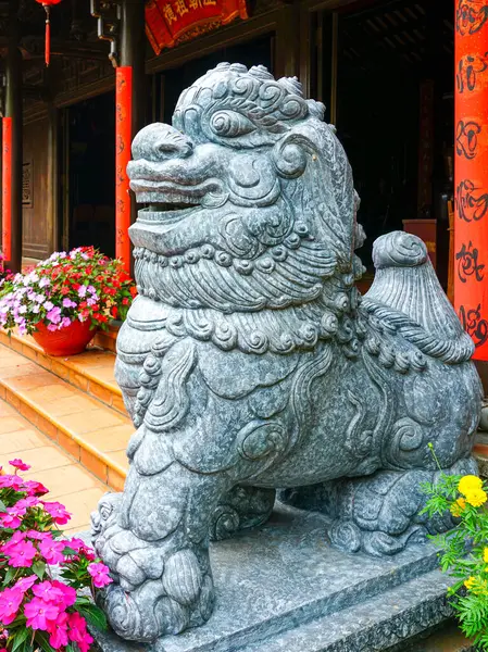 Geleneksel taş koruyucu aslan heykeli, renkli çiçekler ve kırmızı sütunlarla Vietnam tapınağının önünde.