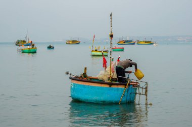 Vietnamlı balıkçı sakin kıyı sularındaki renkli teknelerin arasında geleneksel bir sepet teknesinde çalışıyor, Mui Ne