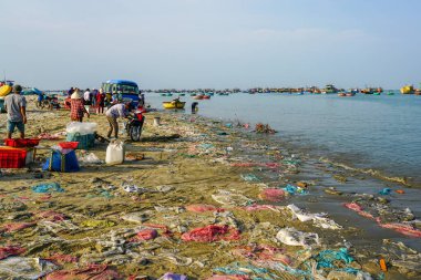 Vietnam balıkçı kasabası Mui Ne kıyıları plastik atıklarla doludur, insanlar toplanır ve arka planda kayıklar yüzer, açık hava balık pazarı