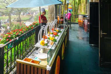 Ba Na Hills, Vietnam- 17 Mart 2025: Bahçe terası büfesi dekoratif şiş tabaklar ve bir aşçı, turistler ve çiçeklerle çevrili