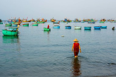 Vietnamlı balıkçı kadın konik şapkalı, renkli Vietnam tekneleri ve balıkçı tekneleri arasında şafak vakti denize giriyor.