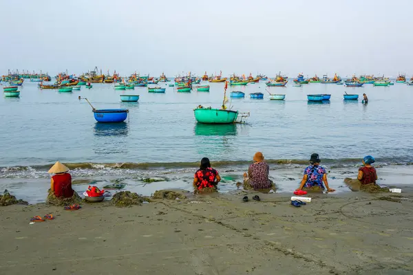 Yerel balıkçı eşleri, huzurlu bir Vietnam plajının kenarında kumların üzerinde oturmuş, sakin denizdeki düzinelerce yuvarlak sepet teknesine bakıyorlardı.