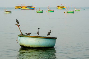 Arka planda renkli balıkçı tekneleri yüzen dört balıkçıl taşıyan yuvarlak bir Vietnam sepeti.