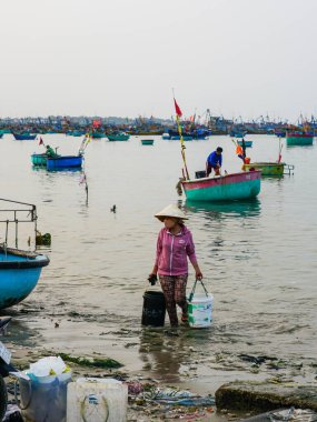 Mui Ne, Vietnam- 22 Mart 2025: Bir kadın, teknelerden kıyıya, deniz çöpleri ve yoğun liman hayatı arasında taze tutulan balıkları plastik kovalarla taşır.