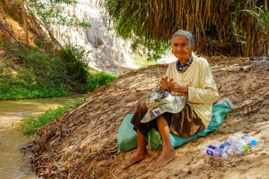 Mui Ne, Vietnam- 22 Mart 2025: Vietnam 'daki Ham Tien Kanyonu' nda çıplak ayakla oturan yaşlı bir kadın, palmiyelerle gölgelenmiş ve yakınlarda akan bir derenin altında kalıyor.