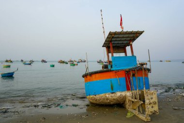 Canlı bir balıkçı teknesi Mui Ne, Vietnam 'da kumsalda dinlenirken, diğer tekneler de sabahın erken saatlerinde sakin denizde yüzüyor.