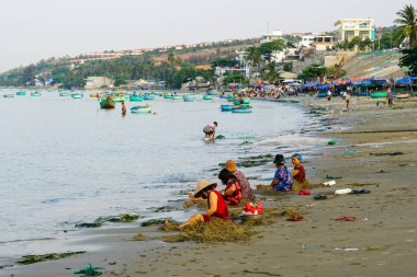 Mui Ne, Vietnam- 22 Mart 2025: Yaşlı kadınlar kumlu kıyı şeridinin tadını çıkarırken geleneksel tekneler sabah güneşinin altında sakince denizde yüzüyor.