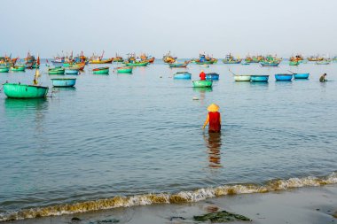 Mui Ne, Vietnam-22 Mart 2025: Konik şapkalı yerli kadın kumsalda kum banyosu yaptıktan sonra denize daldı, huzurlu kıyı sularında deniz tedavisi gördü