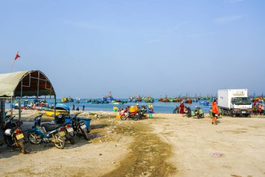 Mui Ne, Vietnam-March22, 2025: Mui Ne limanında balıkçı tekneleri, motorsikletler ve açık mavi gökyüzü altında deniz kenarındaki yerel halkla dolu yoğun bir sabah
