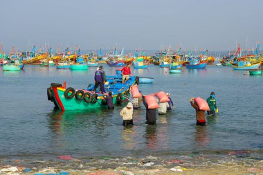 Mui Ne, Vietnam- 22 Mart 2025: İşçiler sığ kıyılarda yük taşıyarak sabah güneşinin altında canlı bir balıkçı teknesine giderler.